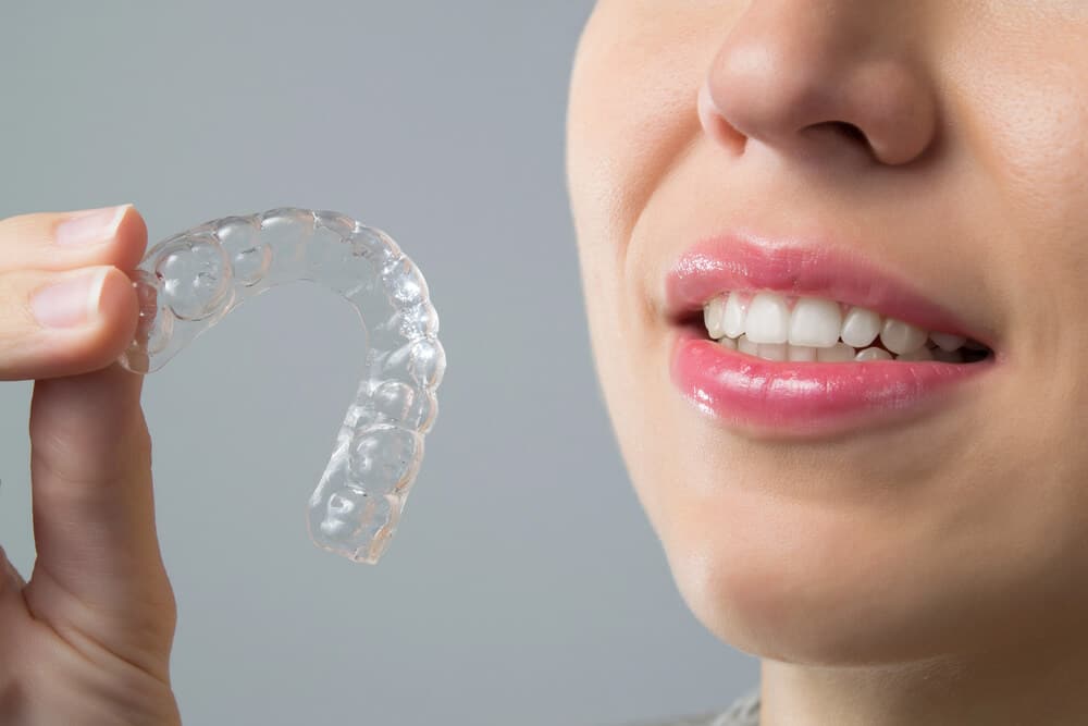 Pacijentica drži prozirni Invisalign aparatić pored svog nasmiješenog lica.